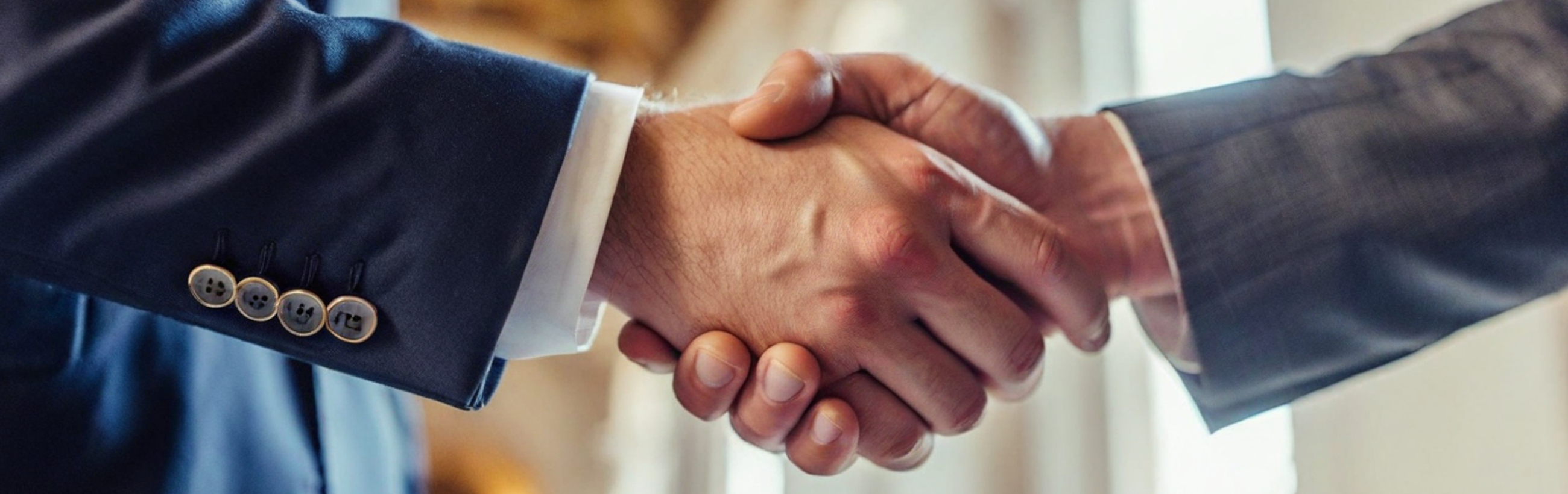 Two men in dark formal suits shaking hands, focusing on their clasped hands, one with visible cufflinks on his sleeve.