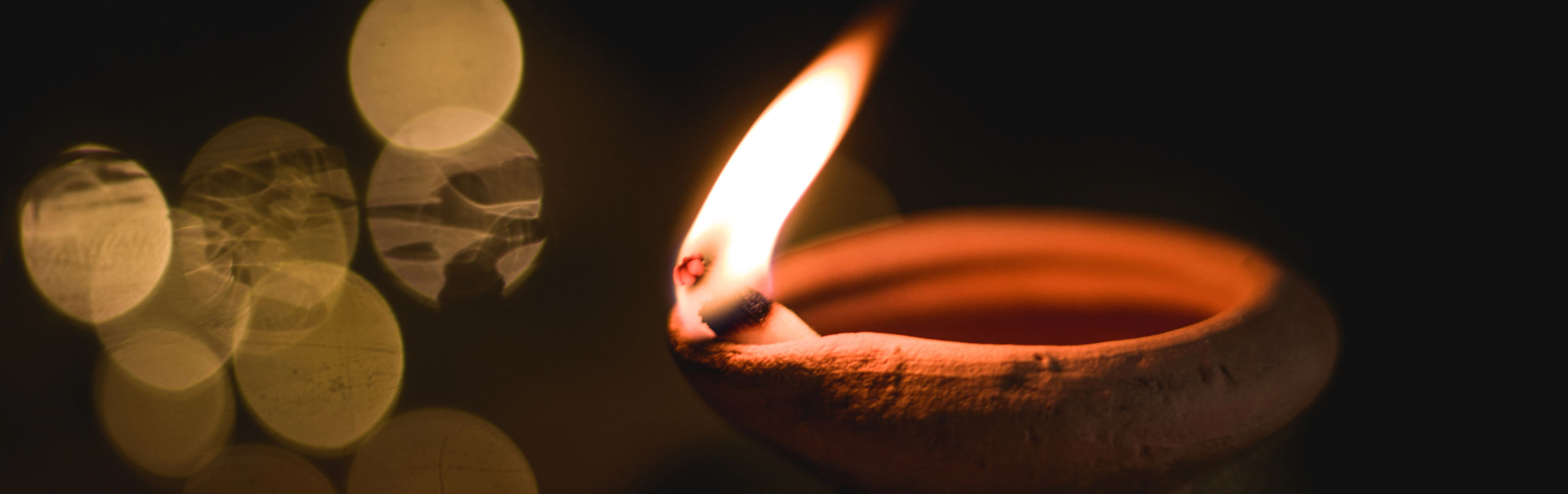 A close-up image capturing the warm glow of a small clay lamp with a single flame prominently lit against a dark backdrop. Subtle light reflections create a soft, bokeh effect mimicking the scattering of faint stars.