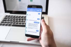 A person holding a smartphone displaying a messaging app, with a laptop with open web pages in the background.