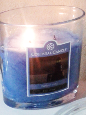 A lit blue candle within a clear glass container, labeled "Colonial Candle Harbor Mist," emitting a soft glow indicative of a serene evening under clear skies.