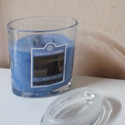 A blue scented candle by Colonial Candle in a clear glass holder with a detached glass lid resting beside it on a white surface.