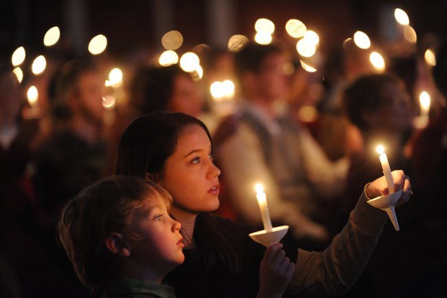 A young woman and a child, attentively participating in a candlelight service, are among a crowd where numerous people hold glowing candles in a dimly lit indoor setting.