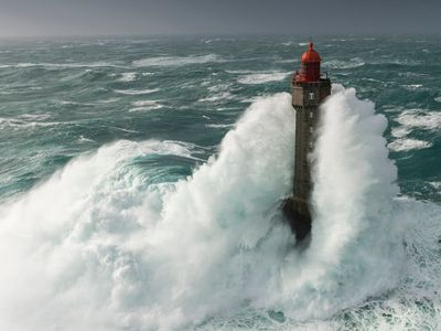 A lighthouse stands resilient against tumultuous ocean waves during a storm, with a fierce wave crashing against it, illuminated by a moody sky.