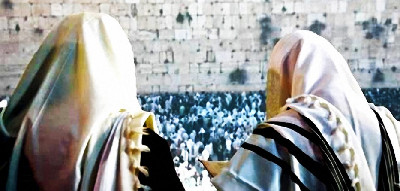 Two individuals wearing traditional prayer shawls look toward a large crowd gathering at a historic stone wall.