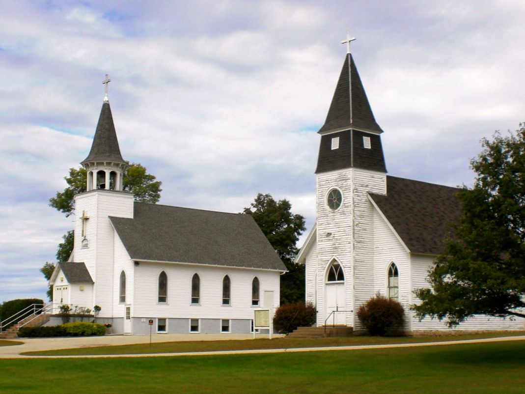 A pair of traditional white church buildings with pointed steeples under a cloudy sky. Each steeple is topped with a metallic weather vane and a cross. The churches feature arched doorways and stained glass windows.