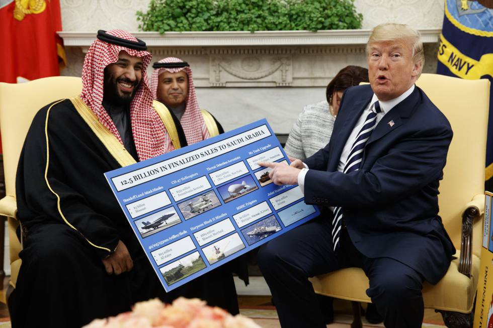 Two men seated in a formal setting with one showing a large board detailing a financial agreement with images of military equipment. The man on the left appears delighted while the man on the right gestures towards the document.
