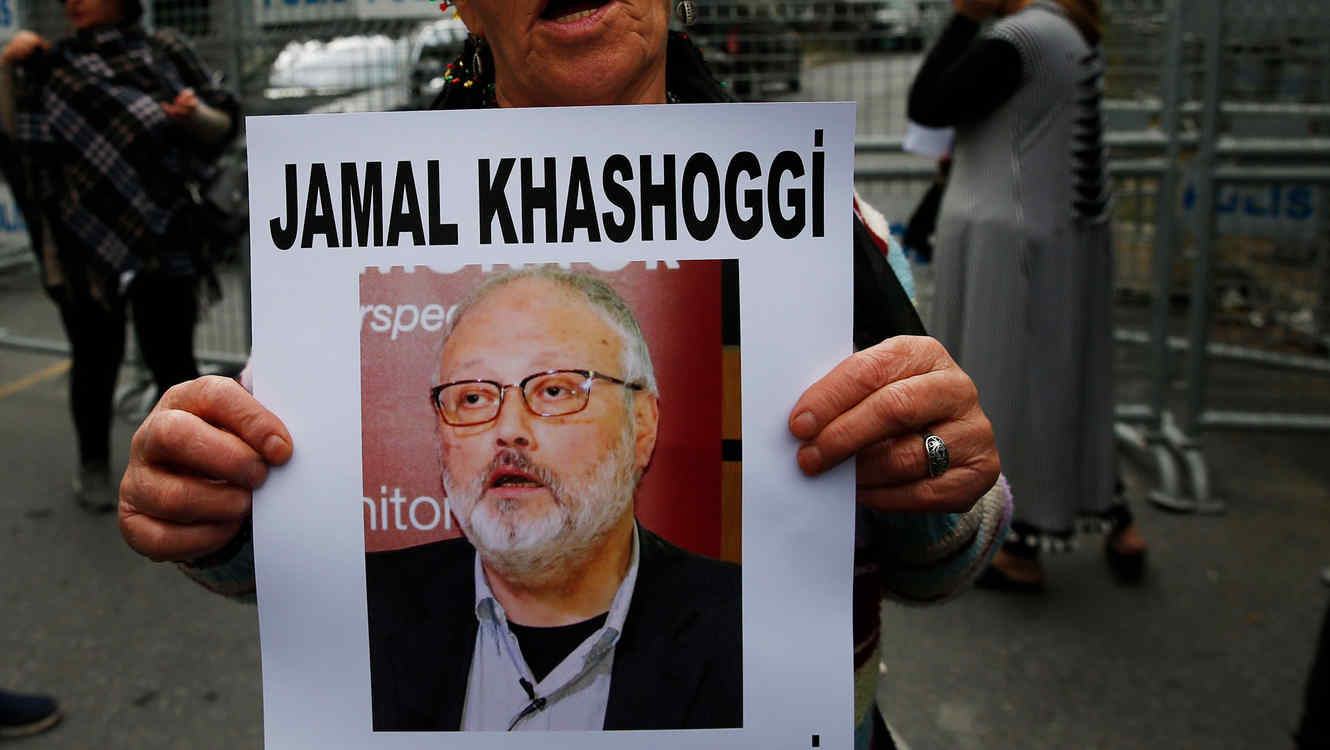 An individual holding a poster of a man with a beard and glasses. The poster features the name "Jamal Khashoggi" and words partly visible that read "perspective" and "monitor".