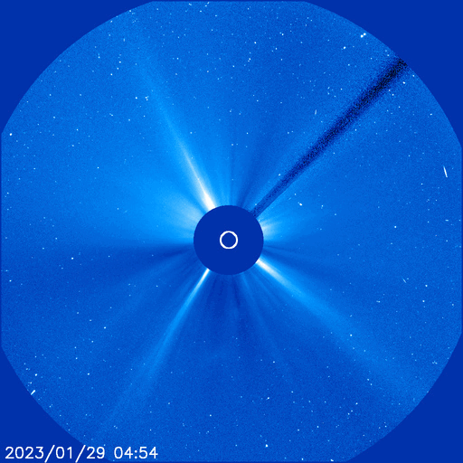 An astronomical image showing the sun blocked by a coronagraph creating a silhouette with radial rays extending outward. A trail resembling a cosmic phenomenon crosses through the celestial field populated with visible stars.