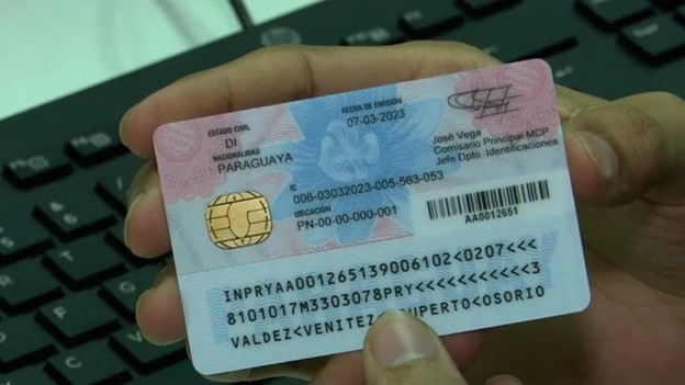 A hand holds a Paraguayan identification card over a laptop keyboard. The card includes personal details, a photo, and security features such as a chip and holographic elements.