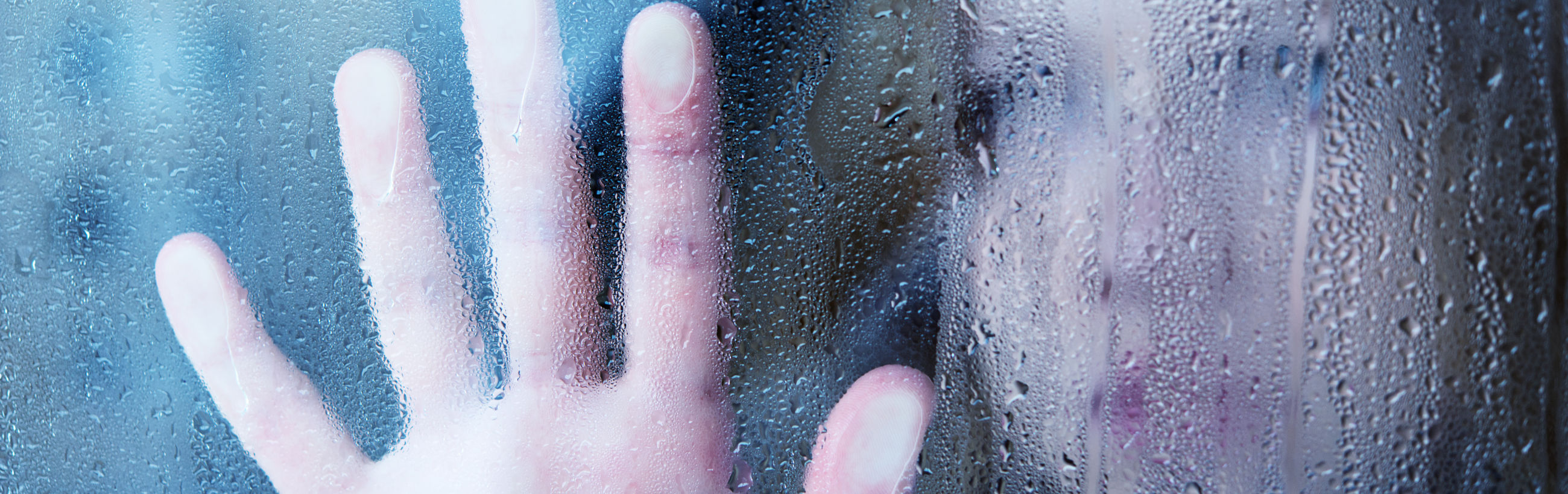 A close-up view of a human hand pressed against a foggy window covered in condensation, distorting the faint silhouette of a face obscured behind the glass.