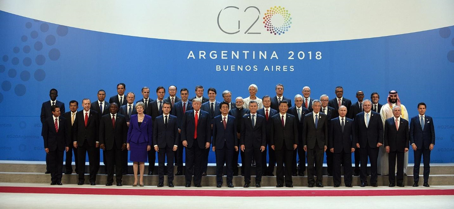 Group of international leaders posing for a photo at the G20 summit in Argentina 2018 against a blue backdrop with event logos and the text "Argentina 2018 Buenos Aires."