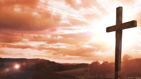 A wooden cross stands prominently in the foreground against a vividly hued sky at sunrise. Rays of sunlight and gentle lens flare grace the serene landscape filled with rolling hills and sparse foliage.