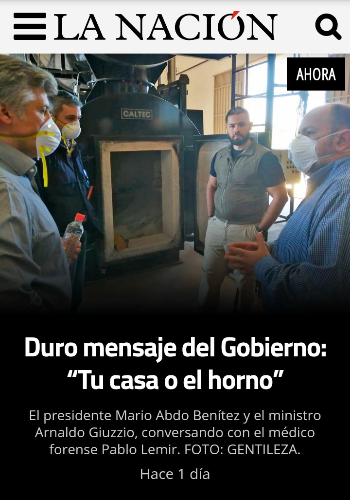 A group of men, including one in a lab coat and others in casual attire, engage in a serious discussion around a large industrial furnace in a workshop setting. The men are wearing protective masks and stand around the furnace that has an open door revealing its interior.
