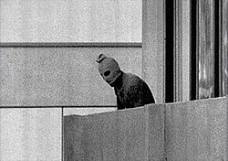A person in a dark suit and alien mask stands on a building ledge, looking out over the edge. The background features a stark, minimalistic building facade.