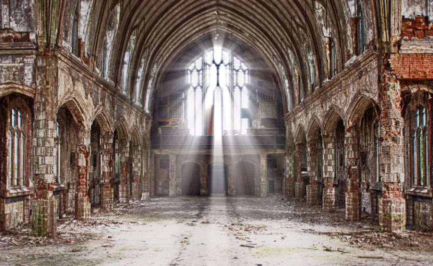 Interior of a dilapidated Gothic cathedral with sunlight streaming through tall, arched stained glass windows illuminating the decaying stone and brick structure, highlighting detailed carvings and the remnants of painted tiles.