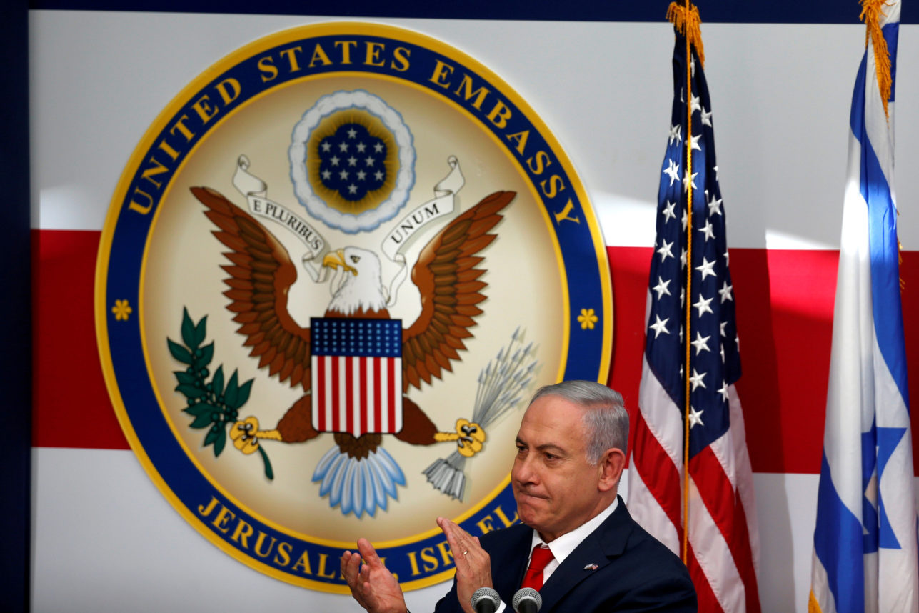 A man stands in front of a large emblem of the United States Embassy in Jerusalem, clapping. The emblem features an eagle with outstretched wings, surrounded by stars and various symbolic elements. The flags of the United States and Israel flank the emblem.