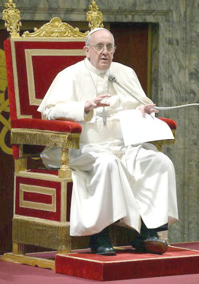 Elderly man in white robes seated on an ornate red and gold throne, speaking and gesturing with one hand while holding papers in the other. He wears glasses and a silver cross pendant.