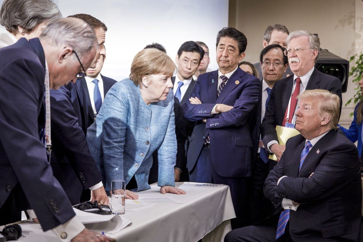 A group of international leaders engage in a discussion around a table draped in white, with maps and documents in sight. Visible are a woman in a blue jacket leaning forward assertively and a man seated in front, sporting a solemn expression.