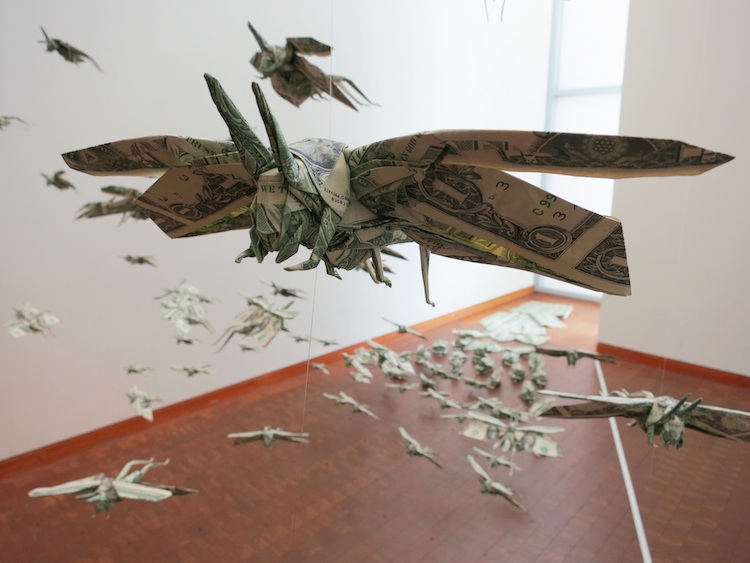 An artistic installation featuring numerous origami models resembling dragonflies, intricately folded from dollar bills, suspended in mid-air within an indoor gallery space, creating an illusion of a flurry or constellation inspired by the Mazzaroth.