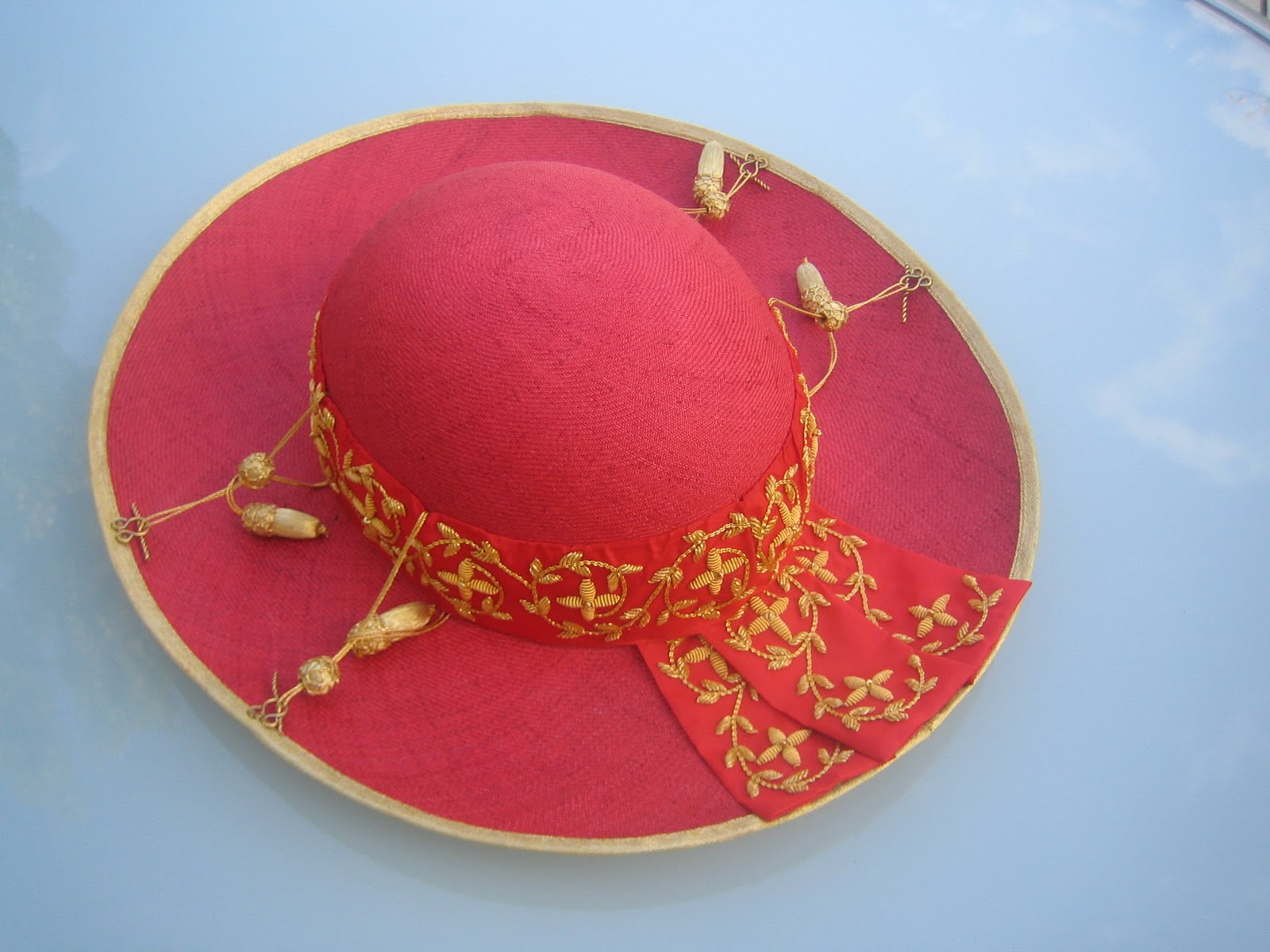 A vibrant red hat with a broad circular brim and a raised dome, adorned with a golden band featuring embroidered patterns. The band is decorated with golden tassels and small spherical accents, set against a reflective blue background.