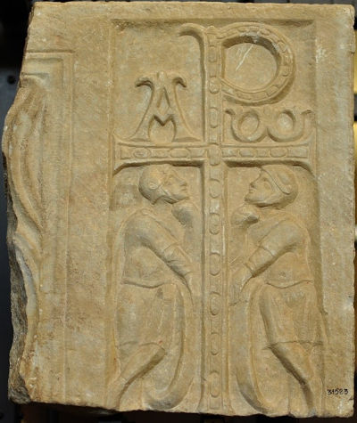 Ancient relief sculpture depicting two human figures facing each other, each placed beside a pillar with intricate patterns and celestial symbols above including a crescent moon. The relief appears to be carved in stone and has historical significance, possibly linked to astronomical themes observed in early cultures.