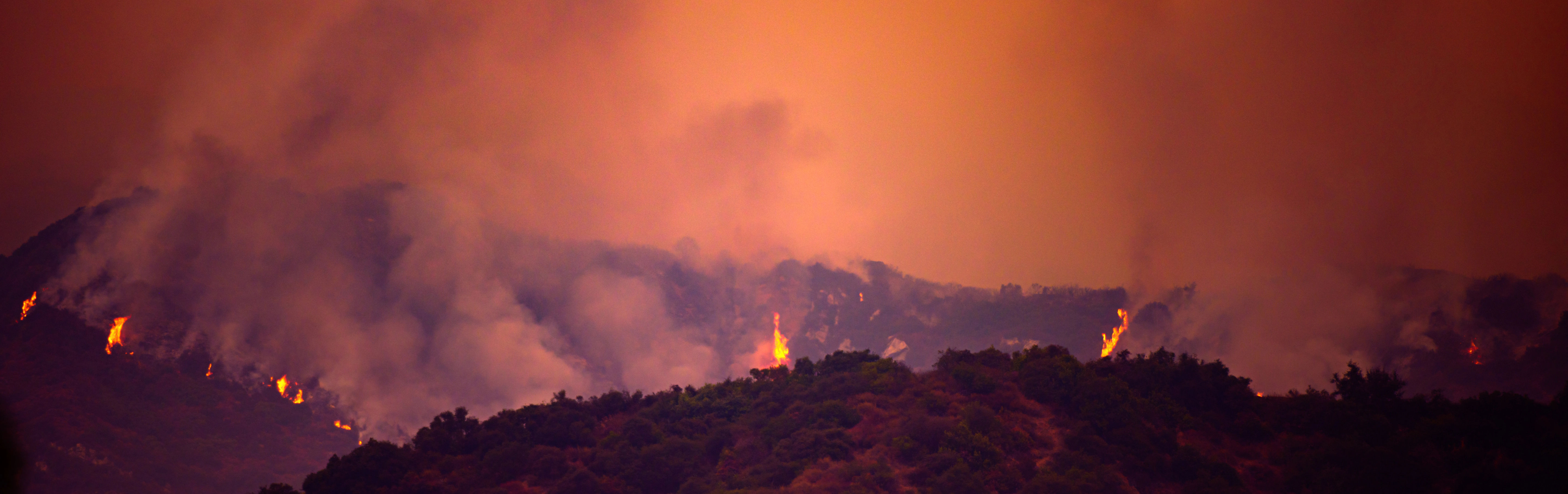A panoramic view of a mountainous landscape engulfed in flames under a heavy, ominous orange sky. Smoke billows across the scene, partially obscuring the fiery peaks where individual flames are visible along the forested ridge.