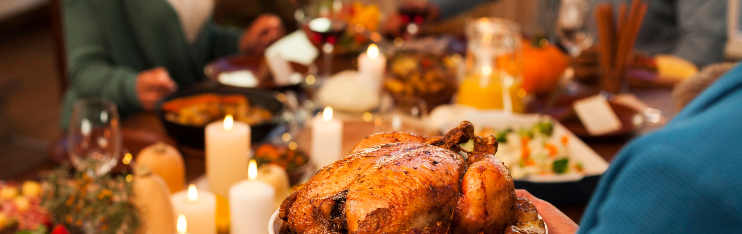 A bountiful dinner scene with a golden-brown roasted chicken at the center surrounded by various dishes, candles, and drinks on a festive table, with blurred figures of people engaged in a meal.