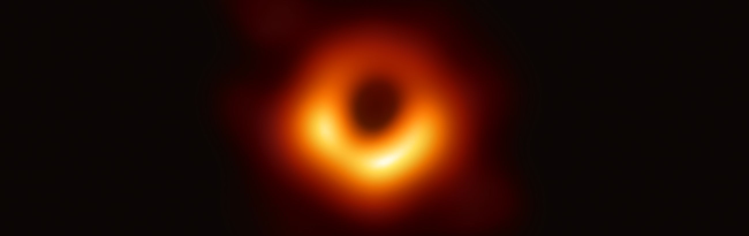 A high-resolution image depicting an astronomical phenomenon showing a central dark area surrounded by a ring of luminous, intense orange and yellow light, gradually fading into the surrounding darkness. Represents a celestial structure in the vast universe.