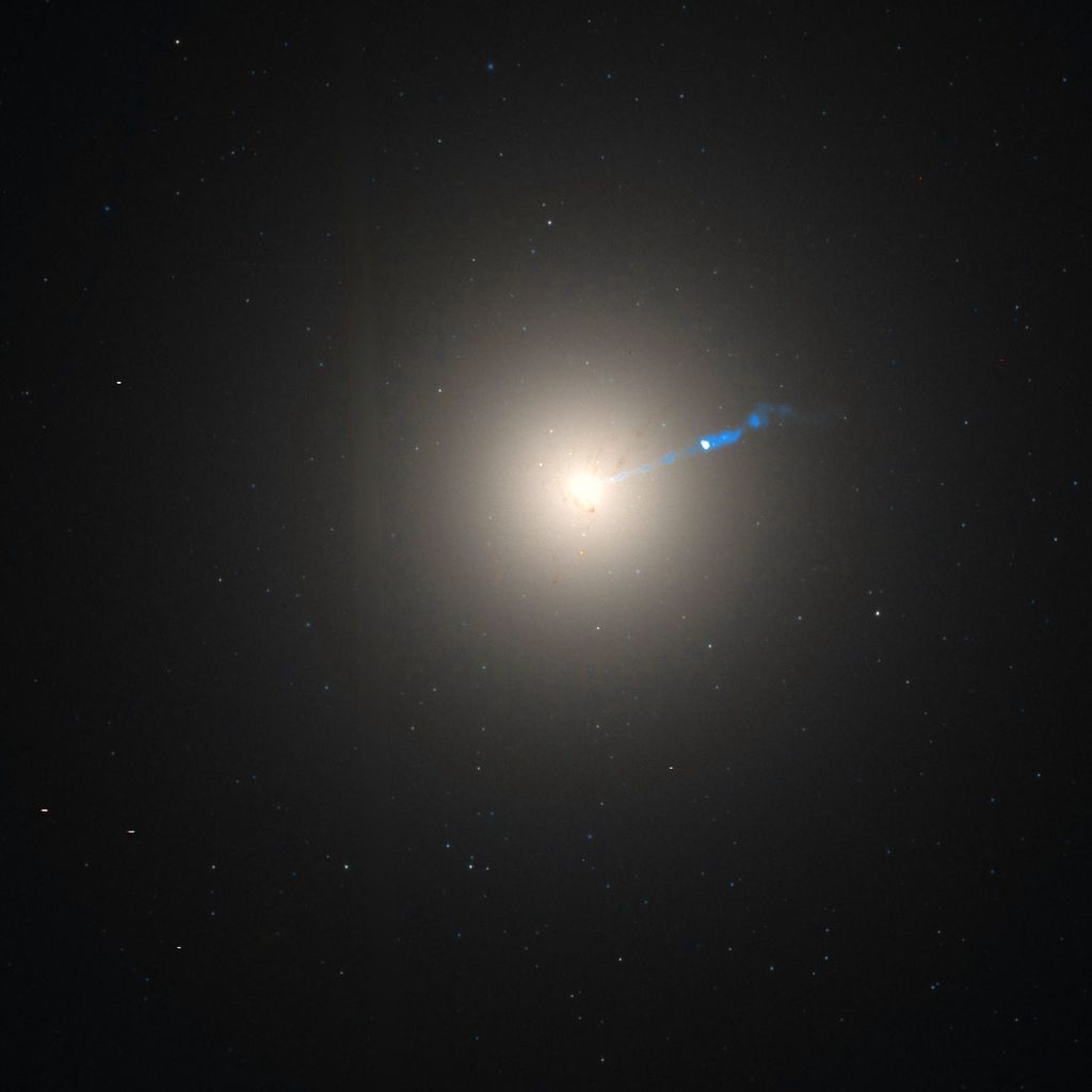 An astronomical photograph displaying a bright galactic nucleus surrounded by a halo, with a distinct celestial jet emanating from it, set against a dark space dotted with stars.