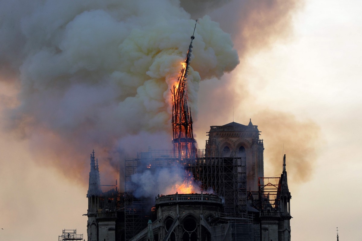 A historic cathedral engulfed in flames and smoke, with its spire dramatically ablaze amidst a thick, billowing cloud of grey and orange smoke against the sky.