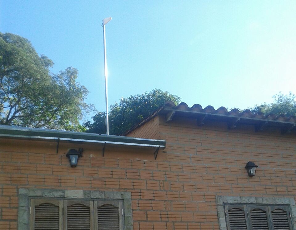 A bright sunny day showcases a house with orange brick walls and typical clay roof tiles. A tall, white pole stands prominently against a clear blue sky, emitting a bright reflective gleam from its top, possibly a solar light. Trees with lush green foliage surround the property, enhancing the serene, suburban atmosphere.