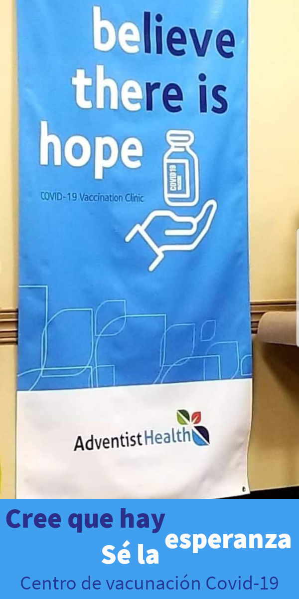 Pancarta promocional azul colgada en una pared con texto blanco que dice "believe there is hope" promoviendo una clínica de vacunación COVID-19. Muestra una ilustración del frasco de vacuna sostenido por una mano y el logo de Adventist Health en la parte inferior.