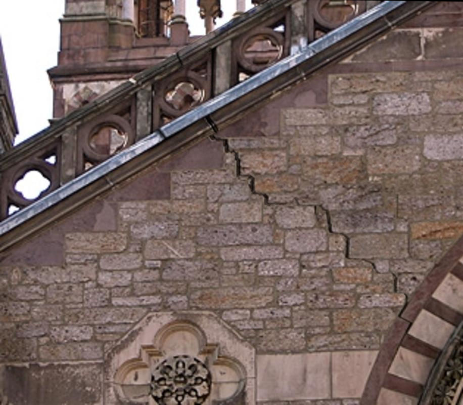 A close-up view of a weathered stone wall featuring a large crack running through it and architectural details including ornate metalwork and patterned openings, all under part of a building's spire.