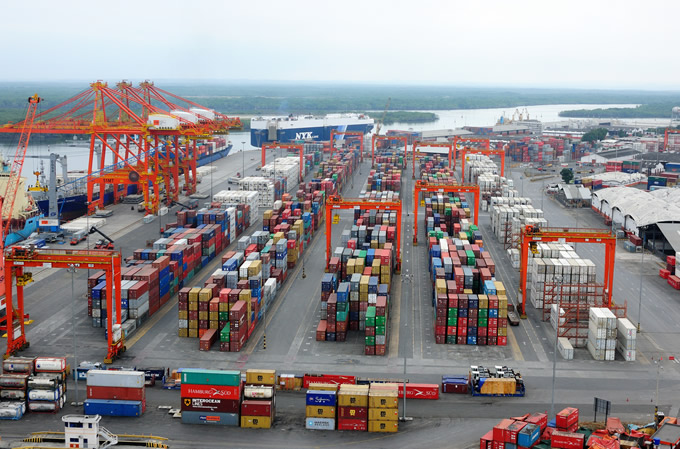 Aerial view of a bustling cargo port showing rows of colorful shipping containers neatly organized and multiple cranes, with a river in the background under a cloudy sky.