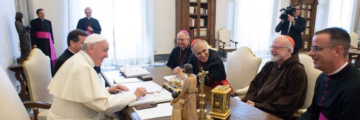 A gathering in a formal office setting with the Pope seated at the center of a long table, engaged in discussion with various clergy members wearing different ecclesiastical attire. A photographer is capturing the moment in the background.