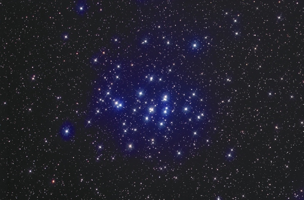 A deep space image showing a dense cluster of stars set against a pitch-black background. The stars vary in size and brightness, creating a shimmering effect across the celestial canvas.