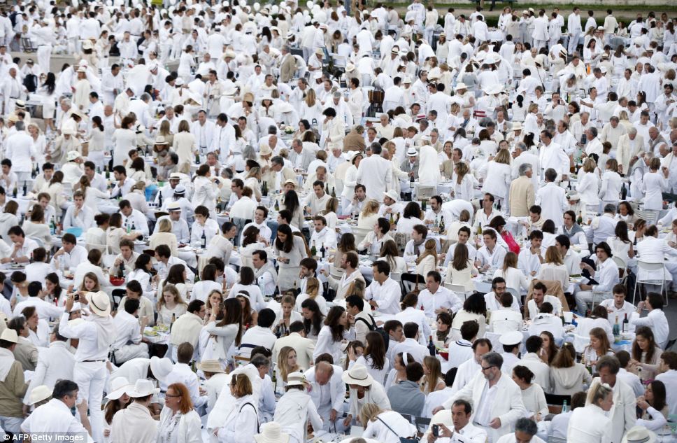A large gathering of people dressed in white attire, seated at multiple tables for a communal outdoor event, evoking the imagery from gatherings depicted in biblical celebrations or feasts.