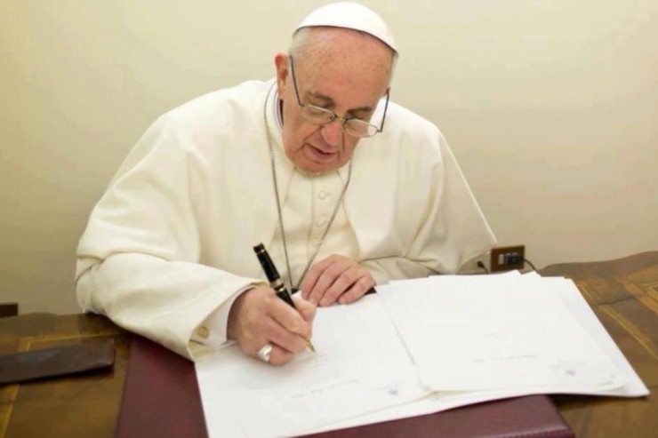 An elderly man in white religious attire is seated at a wooden desk, writing diligently on a document with a black pen. A leather-bound book and additional papers can be seen on the desk.