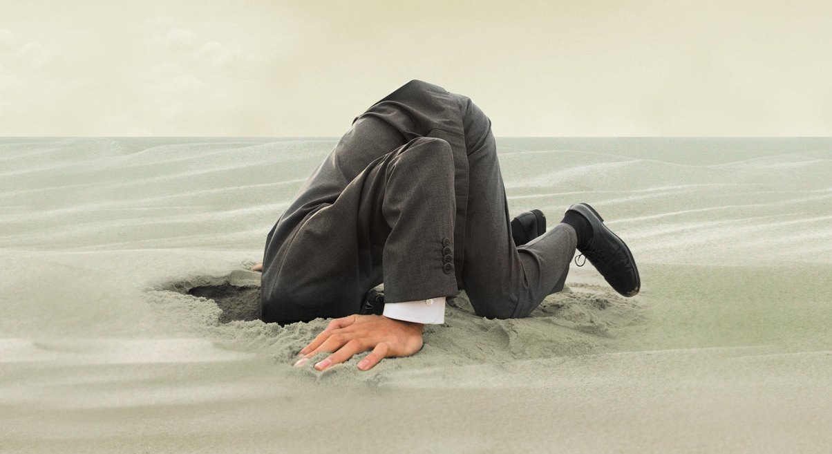 A man dressed in a business suit is depicted with his upper body buried in sand, suggesting concealment or avoidance. His visible hands and feet emerge from the smooth sand, set against a pale, overcast sky.