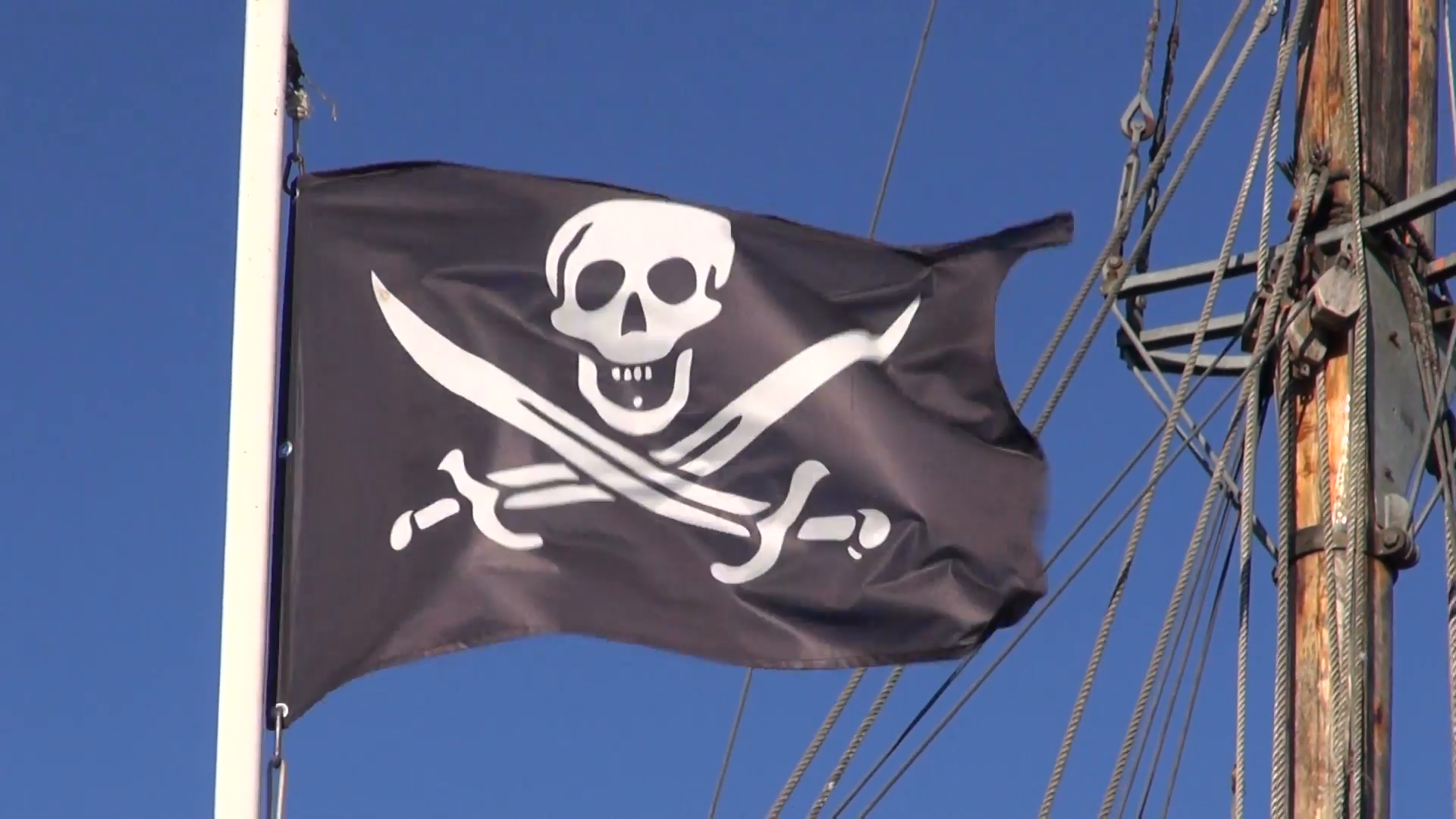 A black pirate flag featuring a white skull and crossed bones flaps against a clear blue sky, attached to a tall ship's mast with ropes and rigging visible in the background.