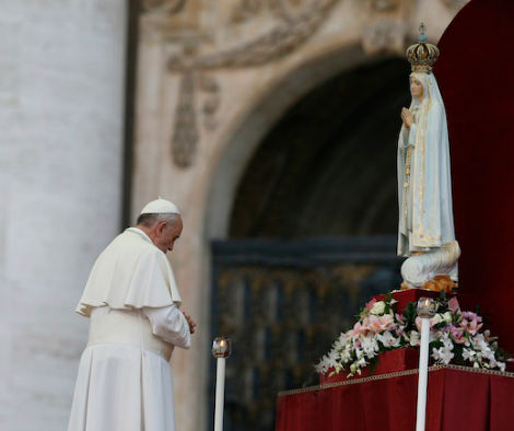 A religious figure in white robes and a white skullcap bows respectfully before a statue depicting a figure in a long blue and white robe, crowned and holding a scepter, standing on a globe and serpent atop a red draped pedestal inside a historic building with an ornate arch.