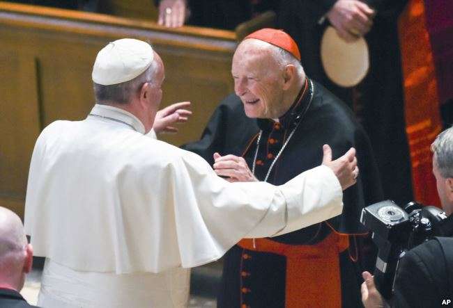 Two elderly men, one dressed in white and the other in red and black clerical attire, joyfully embrace during a public gathering, surrounded by onlookers and a photographer capturing the moment.