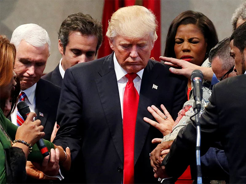 A group of people holding hands and praying around a man in a red tie bowing his head somberly, conveying a scene of collective invocation or intercession.