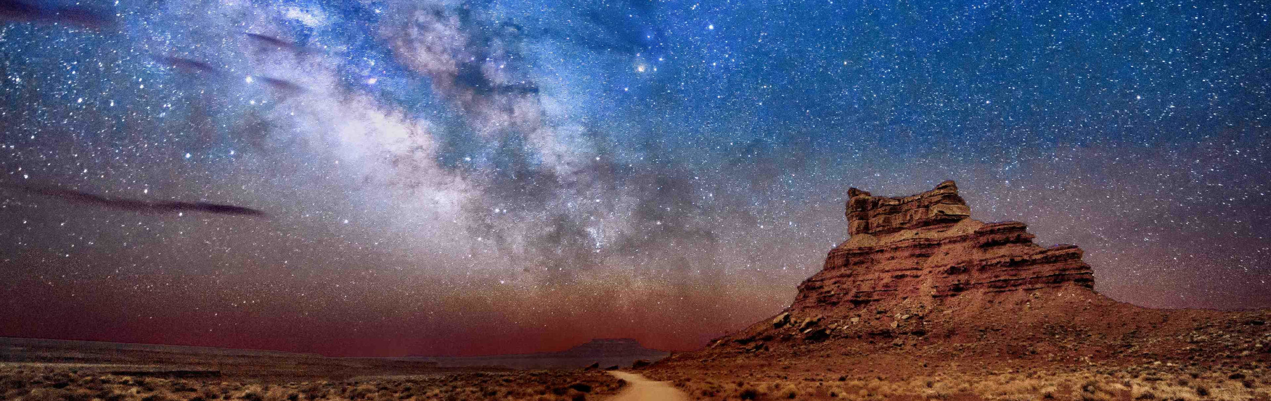 Panoramic desert landscape at night under a vibrant star-filled sky, featuring a towering stratified rock formation on the right. A dusty trail fades into the distance, enhancing the image's sense of wilderness.