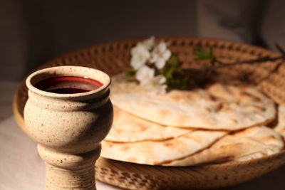 A traditional clay chalice filled with red wine, placed at the forefront on a woven basket that also holds several pieces of unleavened flatbread, with a small bouquet of white flowers adding freshness to the scene. The setting is gently illuminated by natural light, suggesting a serene and historical atmosphere.