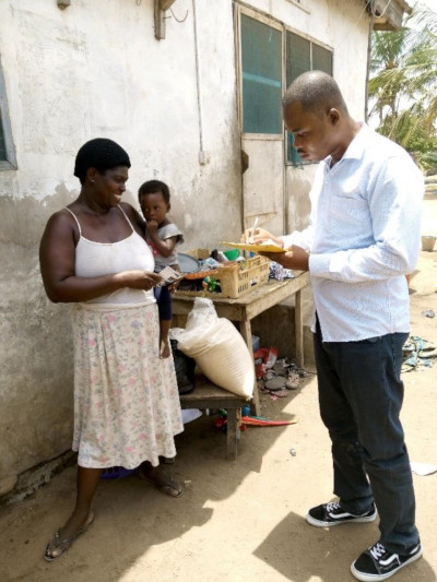 A woman and a man engage in a transaction at an outdoor market stall, with a young child peeking from behind the woman. The woman, wearing a sleeveless top and flower-patterned skirt, appears to be selling goods, while the man, in a light blue shirt and jeans, examines a small item. A humble building and a simple outdoor setting suggest a marketplace in a tropical rural area.