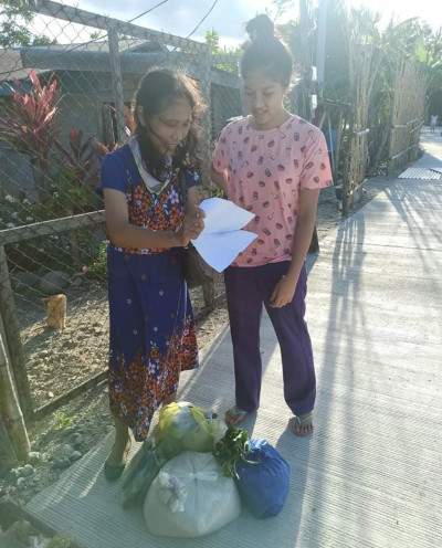 Two young girls standing on a pathway outside, surrounded by foliage, examining a sheet of paper together. One girl is dressed in a floral blue dress and the other in a pink shirt with icons and purple pants. Nearby, several bags full of groceries rest on the ground.