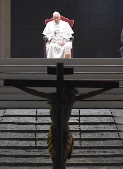A man in white robes and white zucchetto sits on a red chair positioned on a raised stage outdoors, viewed from behind a metal barrier and wooden crucifix in the foreground.