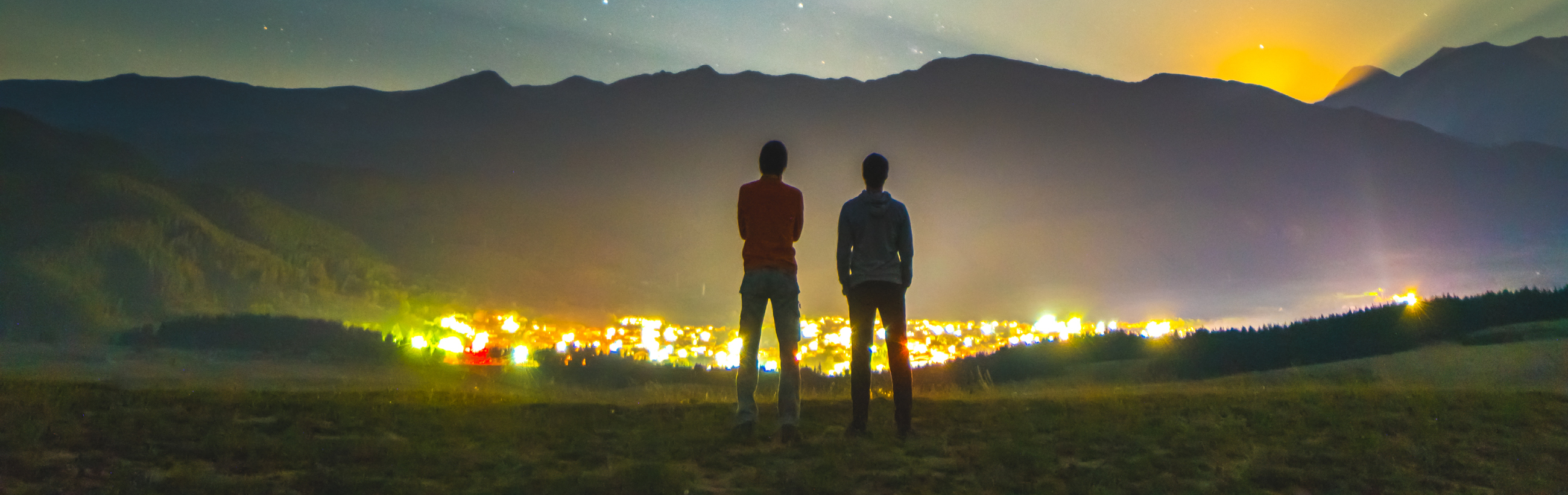 Two individuals standing on a grassy field at twilight, observing the luminous night sky filled with stars and the Mazzaroth. In the background, the soft glow of a town illuminates the base of distant mountains.