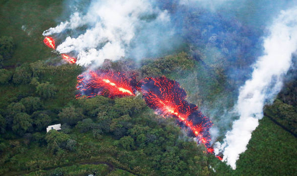 Aerial view of a volcanic eruption with molten lava flowing through dense vegetation, producing clouds of smoke.